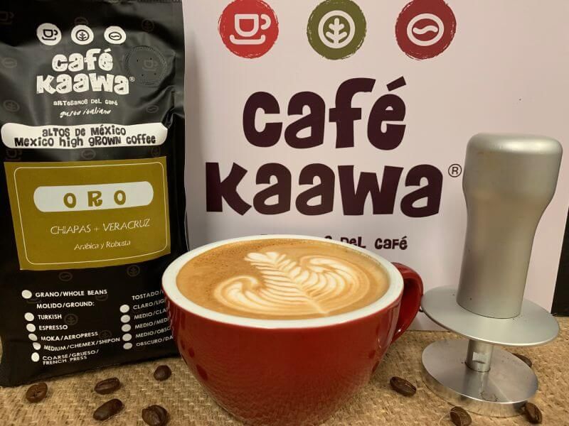 On the back, a Café Kaawa sign and a bag of Café Kaawa Oro. In front of it, there's a red cup of coffee with a tulip pattern on the foam, surrounded by a tamper and coffee beans