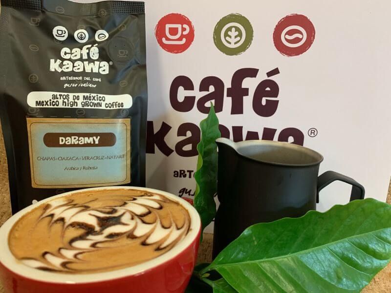 Cup of coffee with intricate latte art design, decorated by surrounding green leafs, a Café Kaawa sign, and a bag of Café Kaawa Daramy